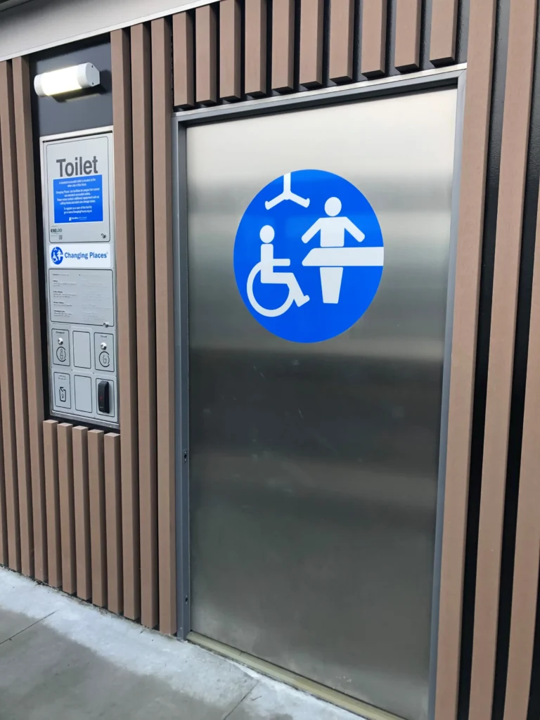The exterior of an accessible public restroom, featuring a large stainless steel door and light brown slatted paneling. A circular blue sign on the door features the universal Changing Places symbol, which shows a person in a wheelchair and a caregiver assisting a person on a changing table. A plaque next to the door indicates that it is a "Toilet" and a "Changing Places" facility.