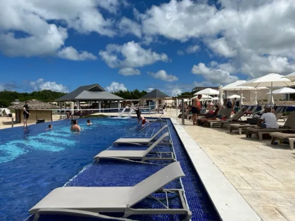 A bright, sunny daytime photo of the main pool area at the Royalton Antigua. The infinity-edge pool features bright blue tile, with submerged lounge chairs lining the edge. Guests are swimming and relaxing on brown lounge chairs under white umbrellas on the tiled deck, with two thatched-roof cabanas and a volleyball net visible in the background against a blue sky with white clouds.