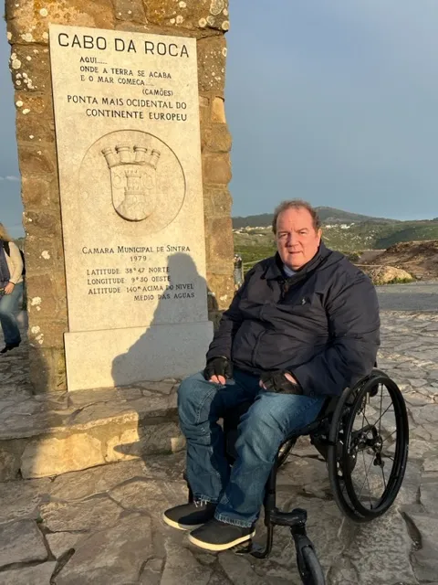 A man in a wheelchair posing next to a stone monument at Cabo da Roca, the westernmost point of continental Europe.