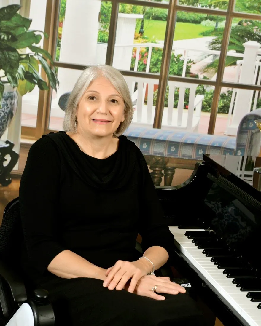 A gray-haired woman in a black top sitting at a grand piano and smiling, with a backdrop depicting a veranda and garden.