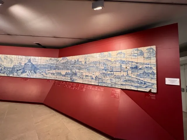 A museum exhibit showing a long, horizontal mural made of blue and white ceramic tiles (azulejos) depicting a historical city skyline, mounted on a red wall.