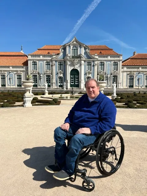 A man in a blue sweater seated in a wheelchair, smiling in front of a grand, ornate European palace with orange roofing.
