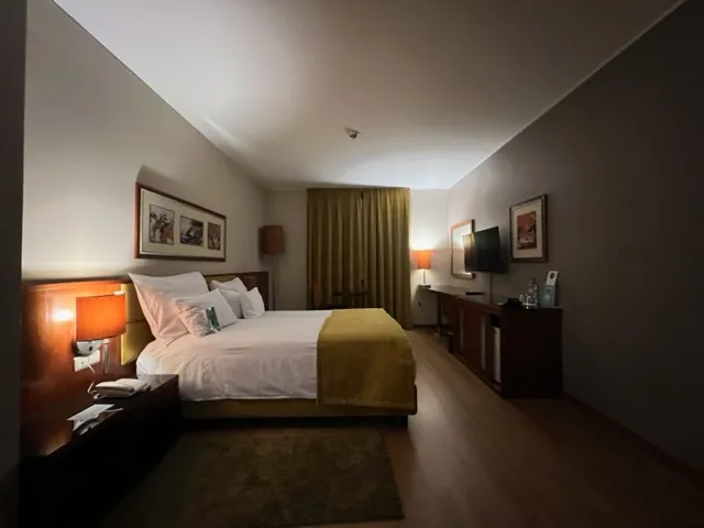 Hotel room interior at night showing a bed with white linens and a yellow throw, with two warm bedside lamps providing soft lighting.
