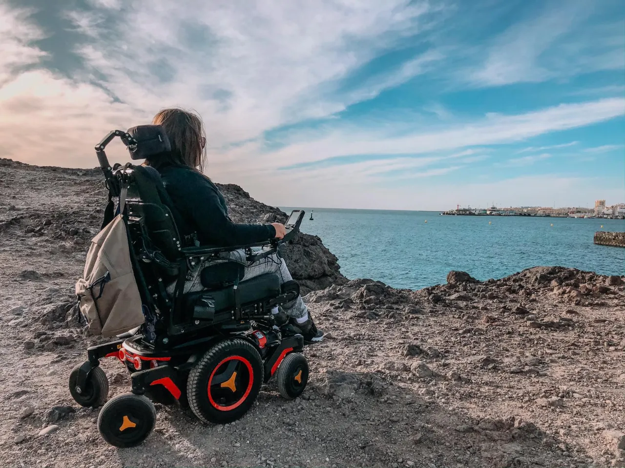 A person is seated in a sophisticated powered wheelchair on a rugged, rocky cliff overlooking a blue ocean. The person is facing away from the camera, looking out towards the sea and a distant port or town on the horizon under a cloudy blue sky.