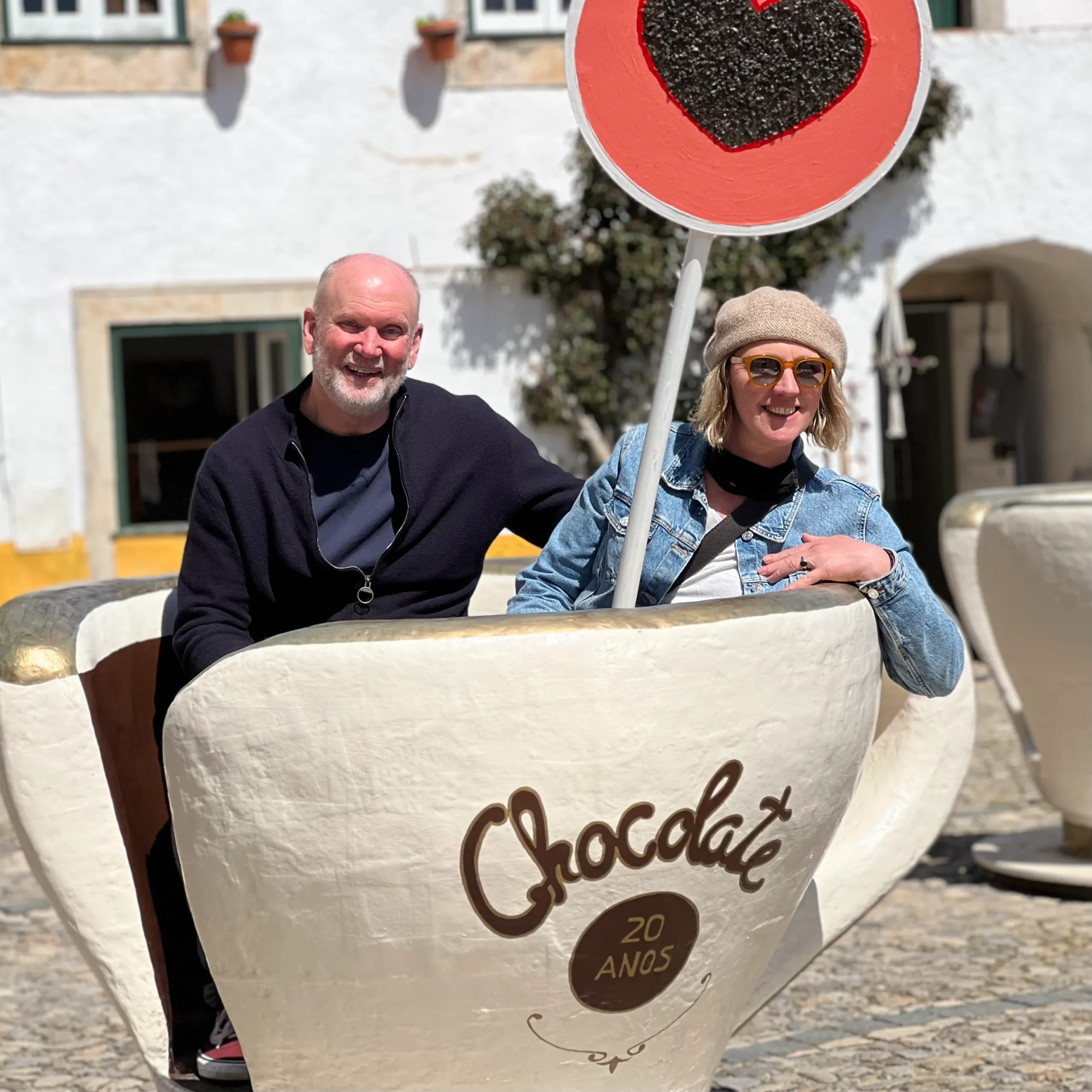 A man and woman are seated inside a very large, decorative white teacup sculpture labeled "Chocolate 20 Anos." The woman is wearing a denim jacket and a beanie, and the man is wearing a black jacket. A sign with a large black heart in a red circle is visible above the cup.