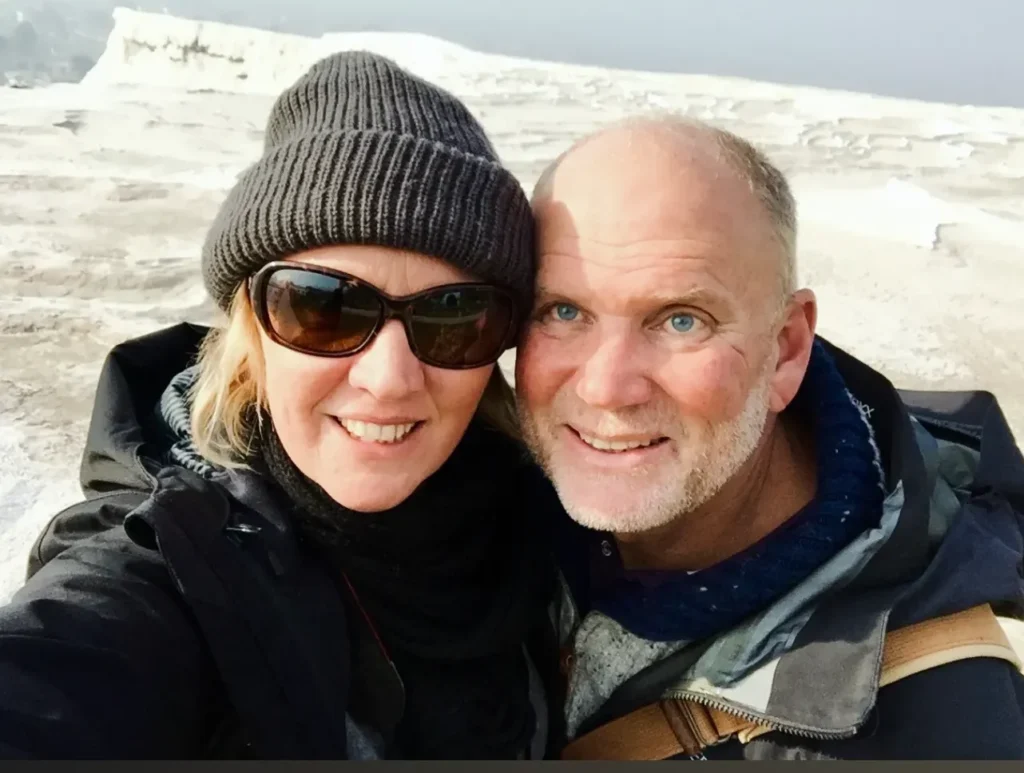 text: A close-up selfie of a smiling man and woman wearing winter clothing and hats. The woman is wearing sunglasses. They are outdoors with a blurry, bright white, rocky terrain visible behind them (likely Pamukkale).