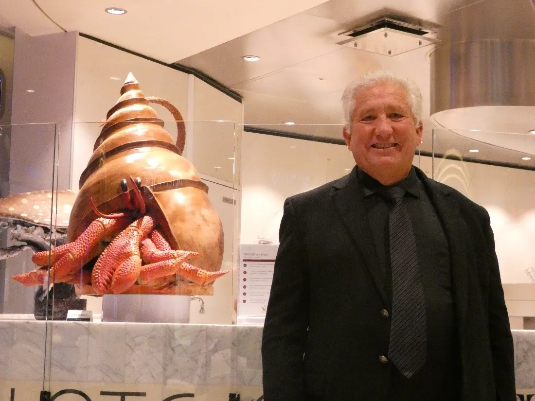 A smiling man in a black suit and tie standing in a modern indoor space next to a large, lifelike sculpture of a hermit crab.