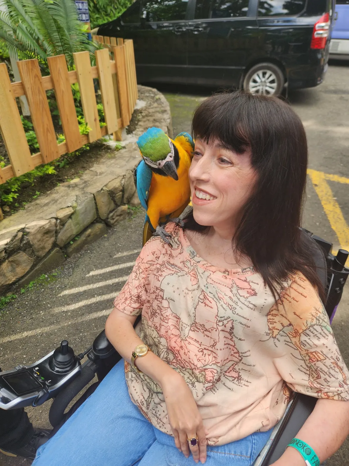 A woman in a wheelchair smiles brightly outdoors while a large blue and yellow macaw parrot is perched on her right shoulder. She is wearing a light-colored t-shirt with a map design and blue jeans.