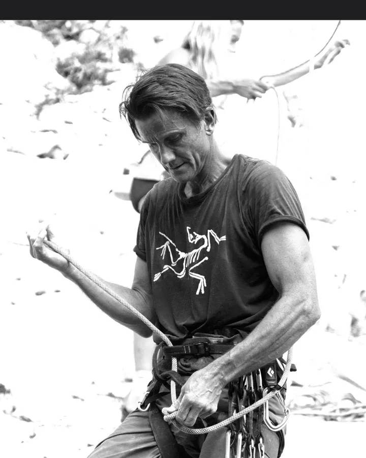 Black and white image of a male climber adjusting the rope and harness on his waist, with another person in the blurred background.