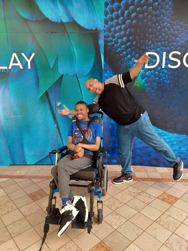 A man standing playfully with his arms outstretched behind a smiling young person who is seated in a wheelchair, in front of a blue feathered wall design.