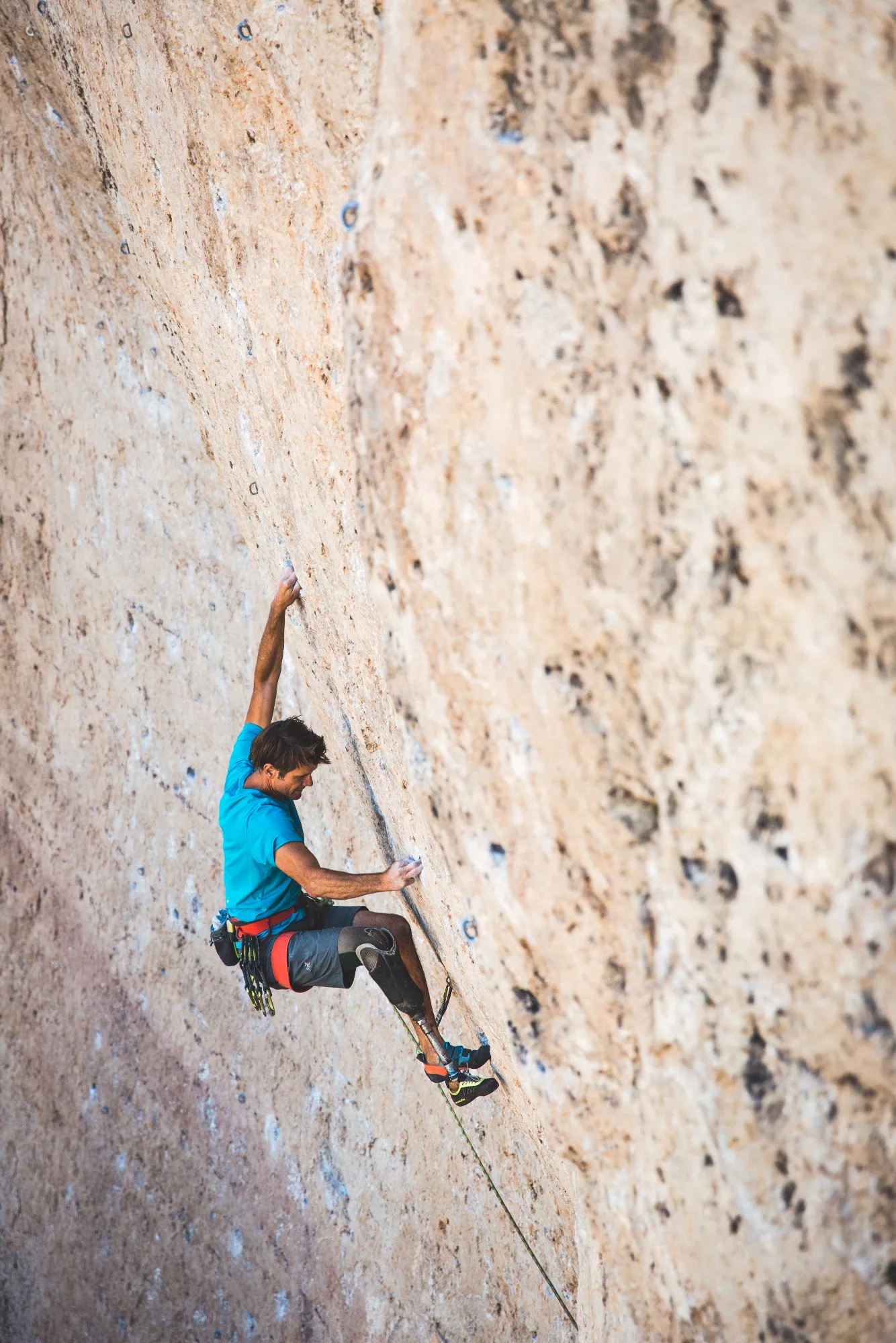 A man wearing a blue shirt and shorts, using a prosthetic lower leg, rock climbing up a sheer, light-colored cliff face.