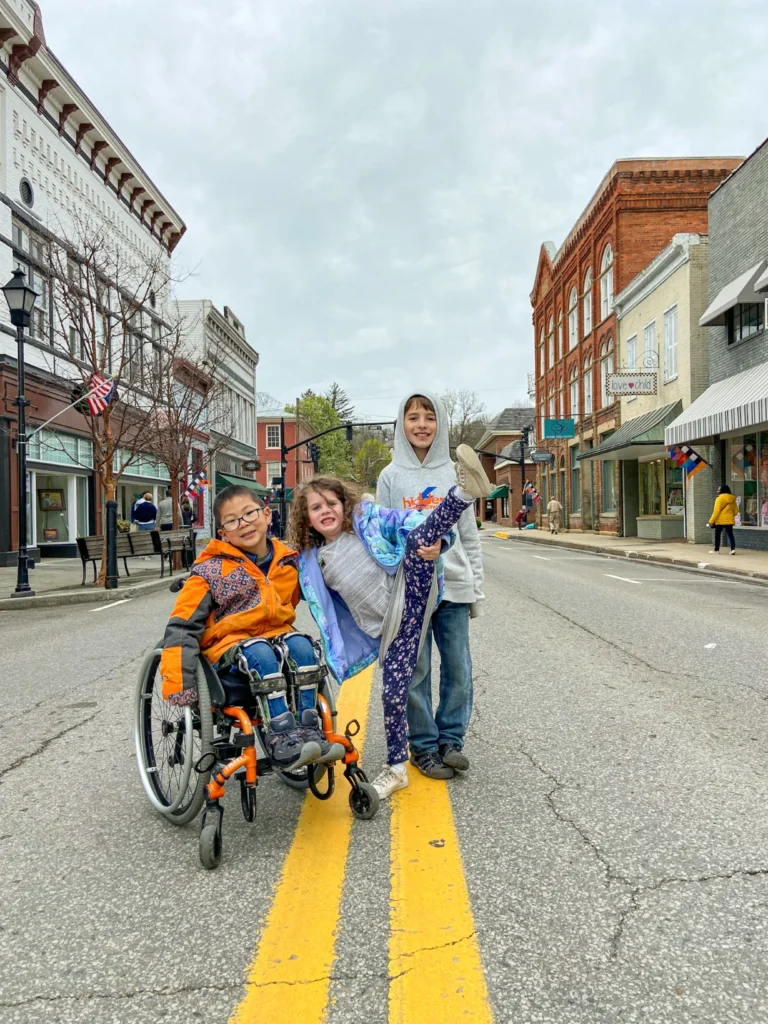 Three smiling children posing on a yellow-striped street in a small historic town; one child is seated in a wheelchair and another child is lifting a leg playfully.