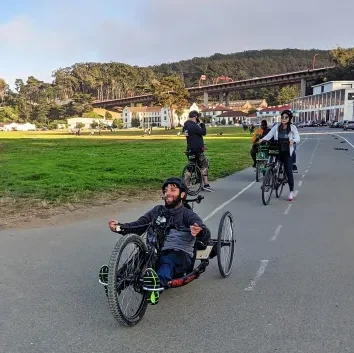 A man smiles while riding a recumbent hand-cycle tricycle on a paved path near a grassy park. Other people are riding bicycles behind him. In the background, there are white buildings and a large bridge (likely the Golden Gate Bridge) crossing a gap between hills.