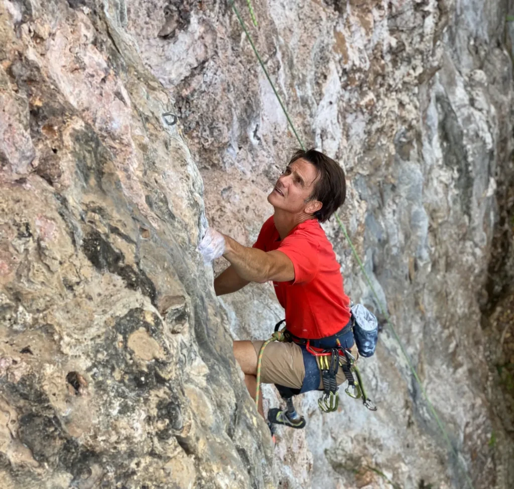 Male rock climber in a red shirt ascending a rough, light-colored cliff, looking up at the route ahead.