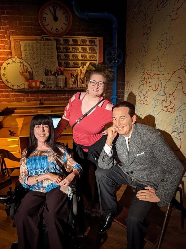 A woman in a wheelchair and another woman standing pose with a wax figure of Walt Disney. They are in a room decorated like an old-fashioned animator's studio with a brick wall, clock, and animation drawings in the background.