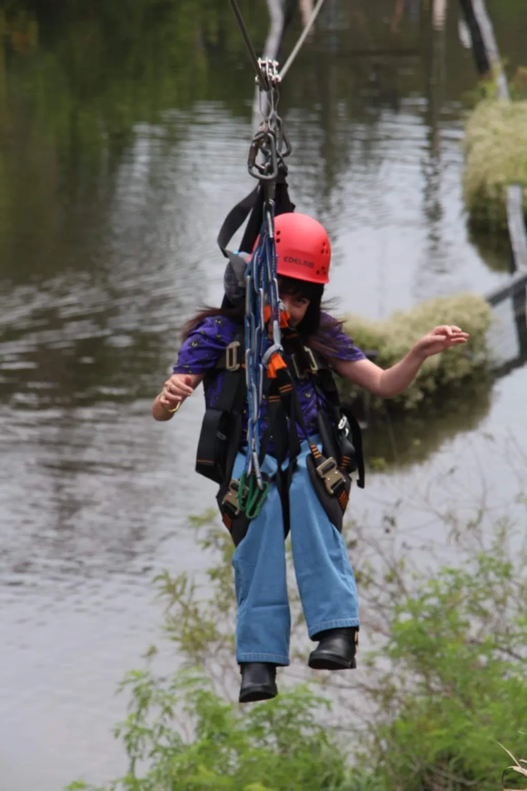 A young person, possibly a girl, wearing a red helmet and blue jeans is suspended by a harness and carabiners while zip-lining over a body of water and lush green foliage.
