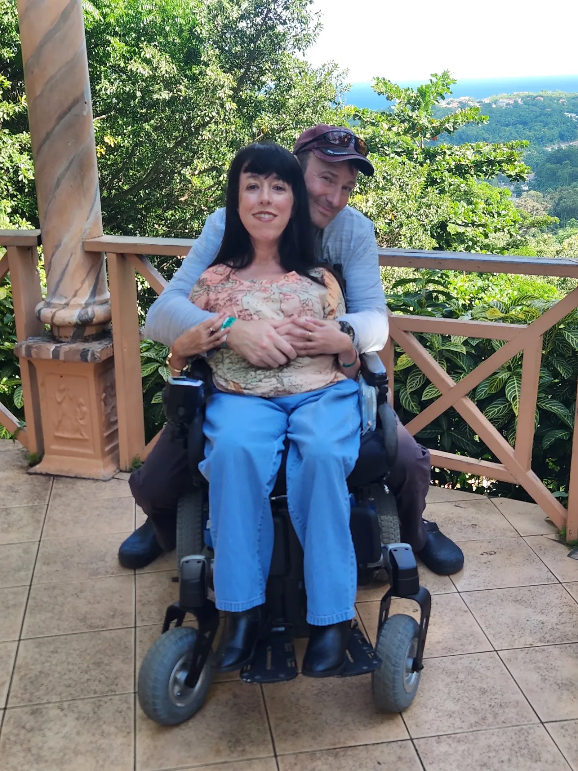 A man is standing behind and hugging a smiling woman seated in a powered wheelchair. They are posing outdoors on a tiled balcony with a wooden railing, overlooking a dense, green tropical landscape that stretches to the ocean/coastline in the distance.