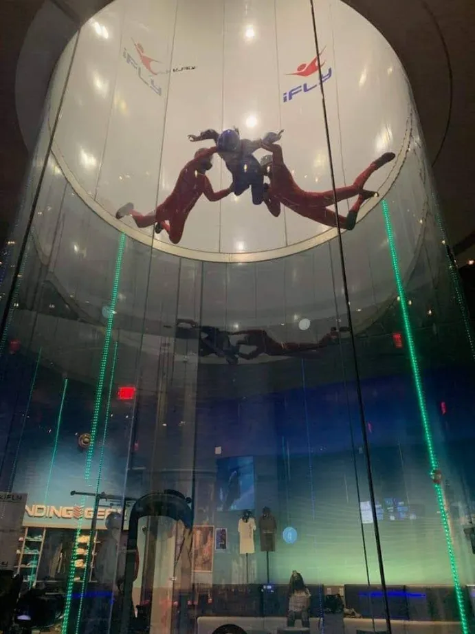 Three people are indoor skydiving inside a large, vertical, clear glass wind tunnel labeled iFLY. They are wearing brightly colored flight suits (two red, one black/dark blue) and are suspended in the air.