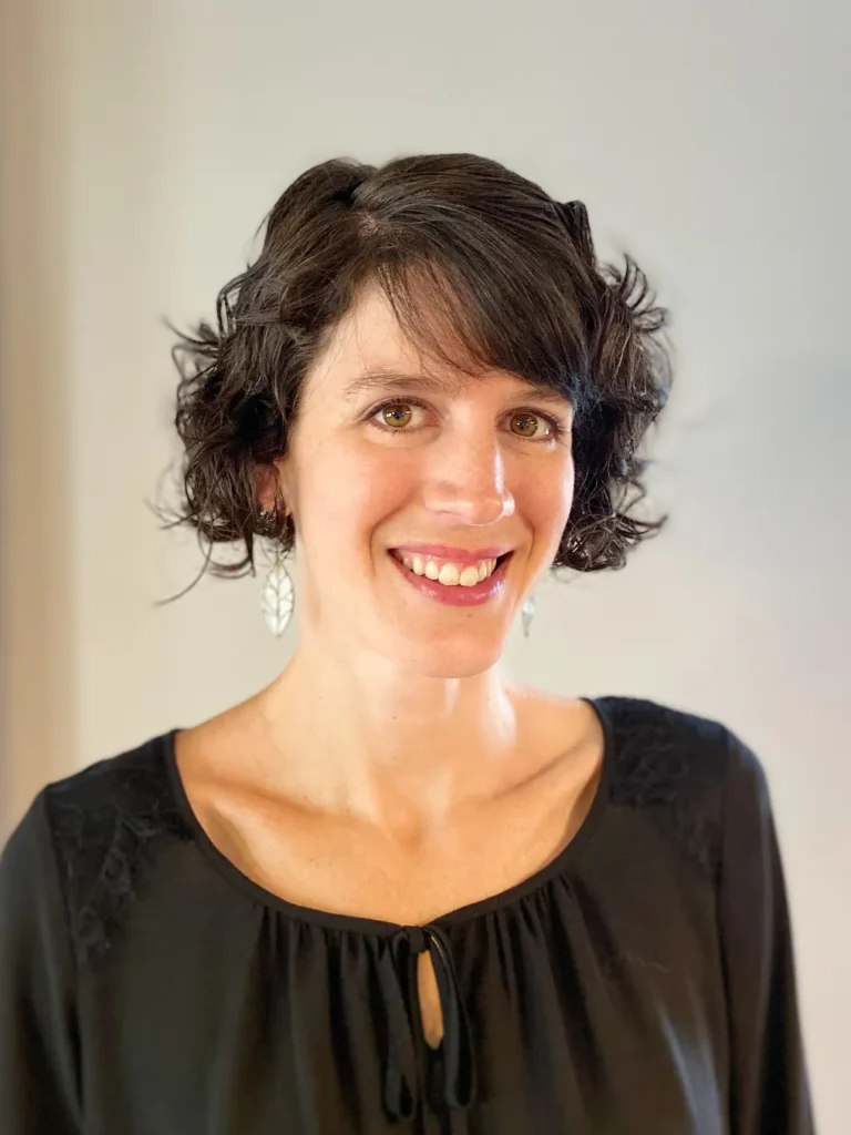 A headshot of a smiling woman with short, dark, wavy hair and brown eyes. She is wearing a black top with lace detailing on the shoulders and long, silver, leaf-shaped earrings. She is standing in front of a neutral-colored wall.