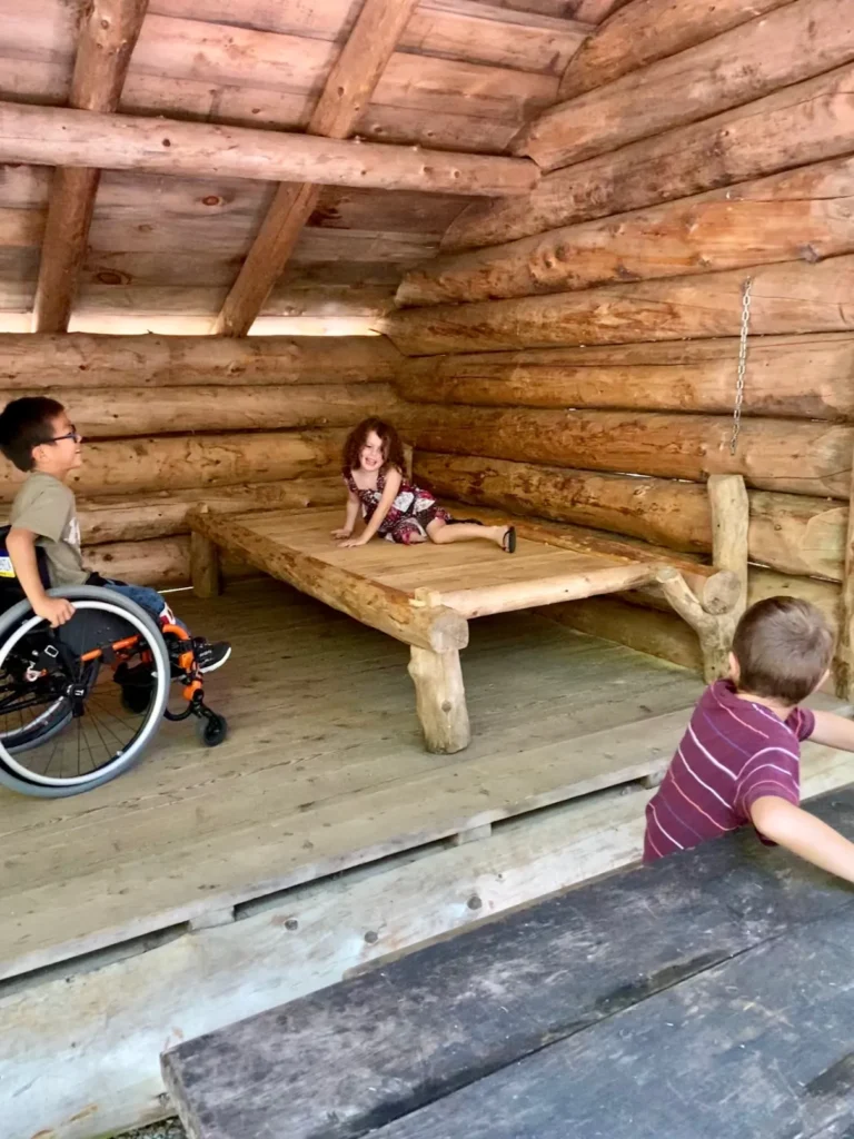 Three children playing inside a rustic wooden log structure with a wooden bed platform; one child is in a wheelchair.