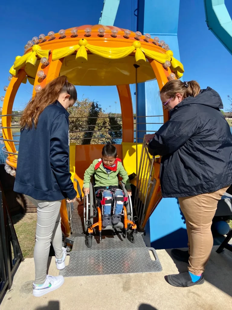Two women helping a young person in a wheelchair roll onto a ramp into an accessible amusement park ride pod on a sunny day.