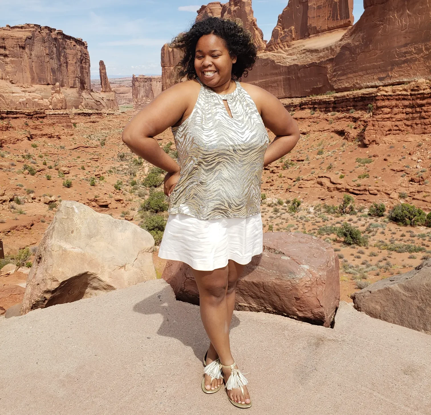 A smiling woman in a shimmery silver top and white skirt posing on a rocky outcrop in a desert landscape with tall red rock formations and canyons.