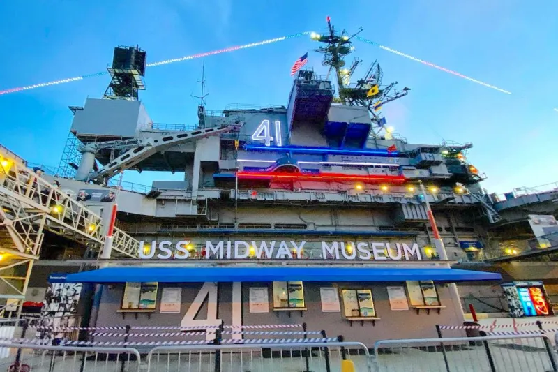 An exterior view of the USS Midway Museum aircraft carrier, docked at a harbor. The ship's hull is marked with the number 41, and a large sign below the flight deck reads "USS MIDWAY MUSEUM" in blue and white lettering. The ship is decorated with colorful lights under a dusky sky.