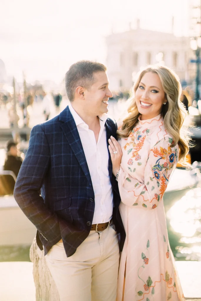 A smiling man in a blue plaid jacket and a woman in a long-sleeved pink dress with colorful floral embroidery posing outdoors in a sunny European city.