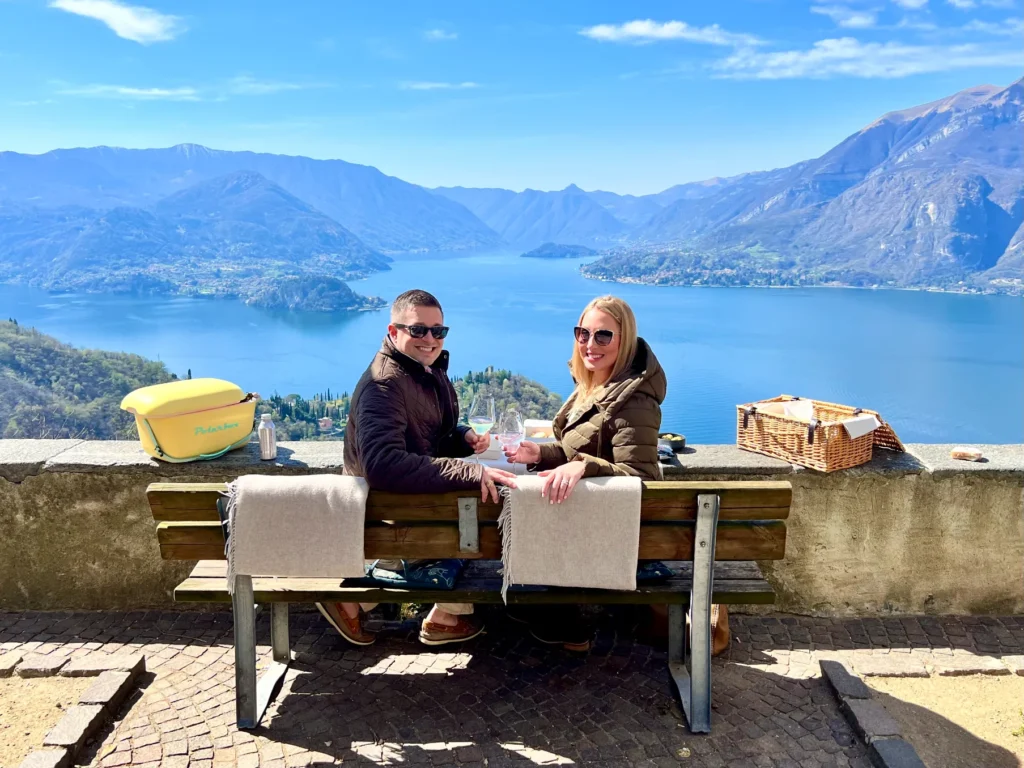 A couple sitting on a wooden bench having a picnic overlooking a large, bright blue lake surrounded by mountains and hills.