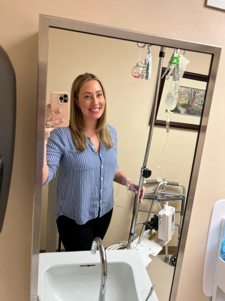 A blonde woman taking a mirror selfie in what appears to be a hospital restroom, smiling while holding an IV pole.