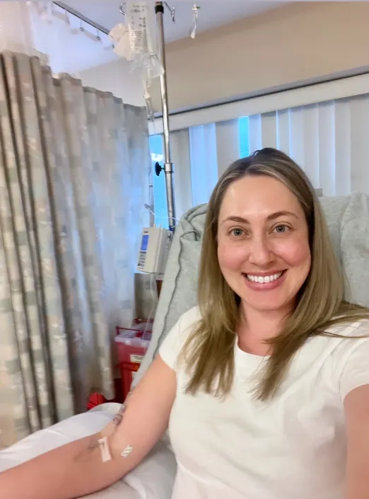 A blonde woman sitting in a hospital bed, smiling at the camera while receiving an intravenous infusion.