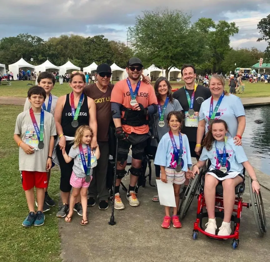 A large, diverse group of ten family members and friends pose outdoors on a path next to a small pond, many of them wearing medals from a race or event. The group includes several children and adults, with two individuals using wheelchairs, and one man wearing a leg brace and using crutches. Several people wear T-shirts with phrases like "I Got Legs" and "GET LEGS." A park setting with large white event tents and trees is visible in the background.