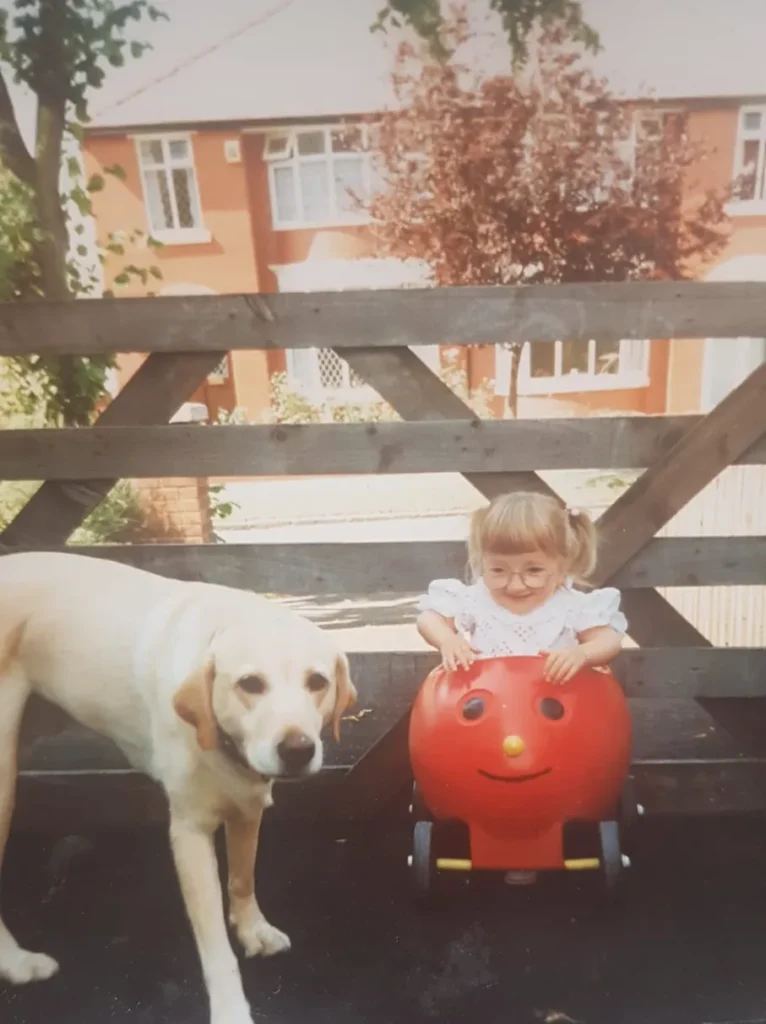 A vintage-style photo of a young toddler with pigtails and round glasses, smiling as she sits behind a wooden fence on a red, apple-shaped toy with a happy face. Standing next to her, on the left, is a yellow Labrador dog looking at the camera. In the background, beyond the fence, a suburban street is visible with houses.