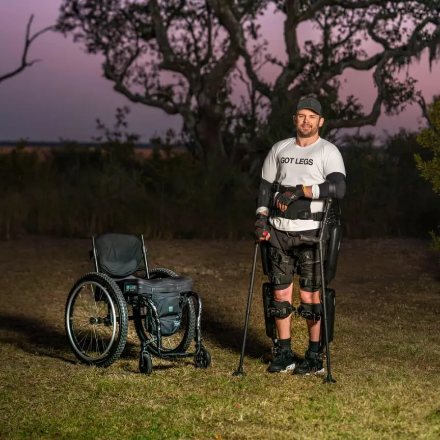 A man stands outdoors at dusk in a grassy field, wearing a white t-shirt that says "I GOT LEGS", a baseball cap, and extensive black exoskeleton leg braces with knee and elbow pads. He is using two crutch-like walking poles for support. To his left, a black wheelchair sits nearby. Behind him are large, leafless trees and a low horizon under a twilight sky.