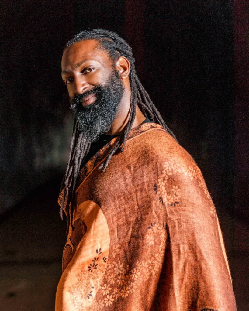 A portrait of a man with long black dreadlocks and a full, thick beard, looking over his shoulder and smiling warmly at the camera. He is wearing a dark top draped with a patterned shawl or wrap in shades of terracotta and orange with a repeating floral design. The background is dark and indistinct, suggesting low, dramatic lighting.