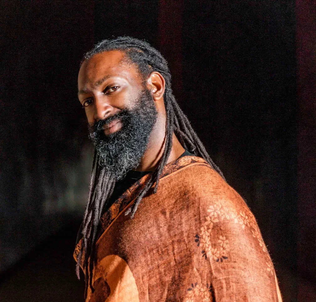 A portrait of a man with long black dreadlocks and a full, thick beard, looking over his shoulder and smiling warmly at the camera. He is wearing a dark top draped with a patterned shawl or wrap in shades of terracotta and orange with a repeating floral design. The background is dark and indistinct, suggesting low, dramatic lighting.