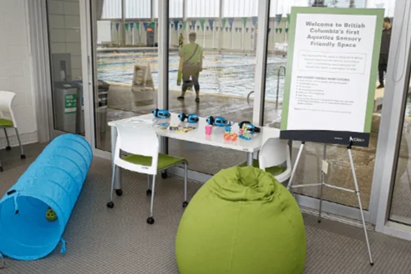 The entrance to an indoor aquatics facility, designated as a sensory friendly space. In the foreground, there is a small table with fidget toys, a large green beanbag chair, and a bright blue fabric tunnel. To the right, a large green sign welcomes visitors to British Columbia's first Aquatics Sensory Friendly Space.