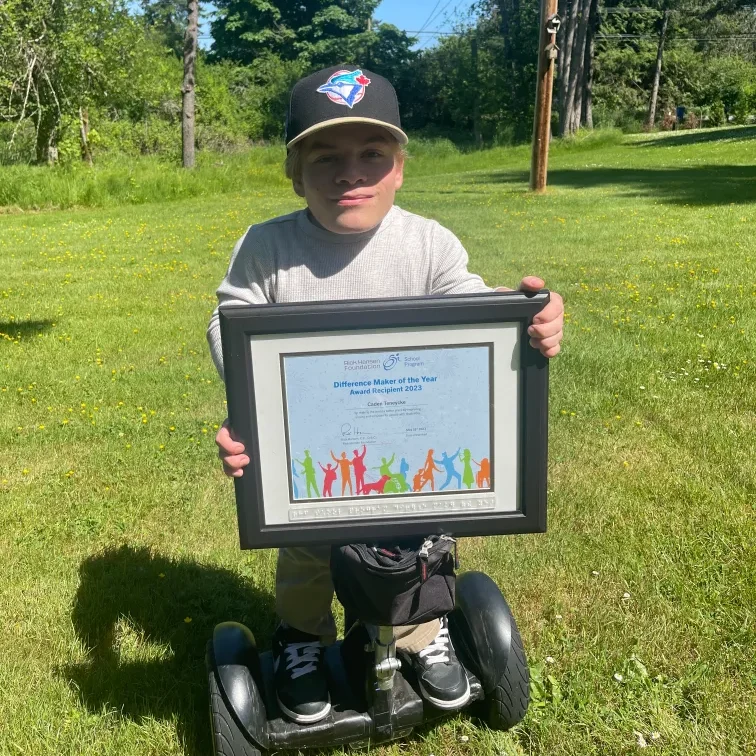 A smiling young boy wearing a Toronto Blue Jays baseball cap and a light grey sweater stands on a black Segway or two-wheeled personal transporter. He is holding a framed certificate that reads "Difference Maker of the Year Award Recipient 2023." He is outdoors on a sunny day in a grassy field with trees in the background.