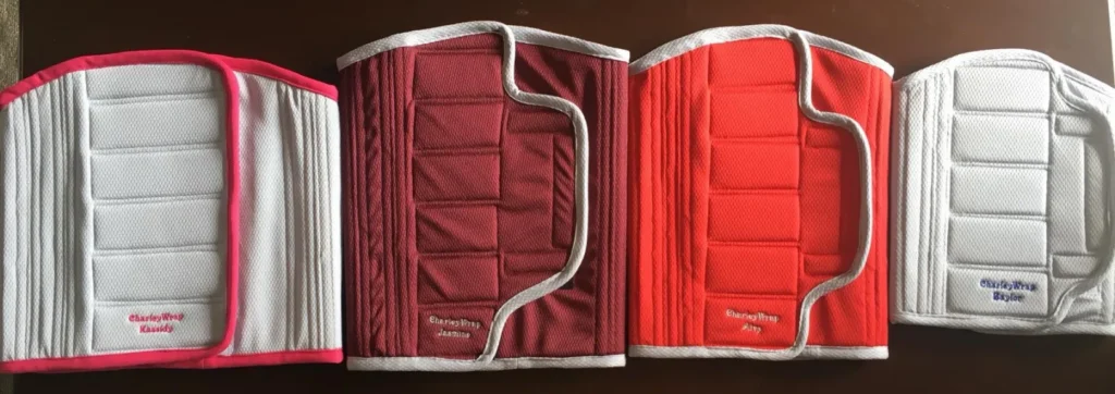 A top-down view of four quilted, rectangular back panels for what appears to be a child's restraint or safety seat, lying side-by-side. Each panel is primarily white and quilted with horizontal rectangles. They are edged and accented in different colors, and each is embroidered with the name "Charley Wrap" and a specific style name: from left to right, "Candy" (pink), "Jasmine" (burgundy), "Roxy" (red), and "Skyler" (grey).