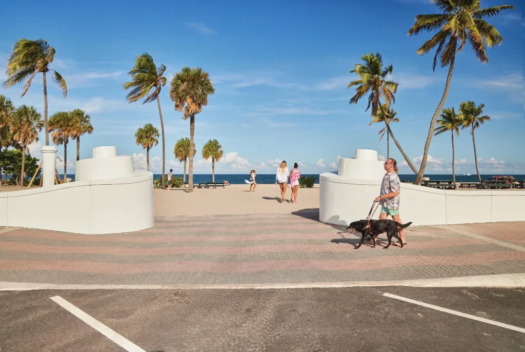 People enjoying a sunny day at Fort Lauderdale Beach promenade with palm trees, blue sky, and ocean in the background, including a blind man with his dog.