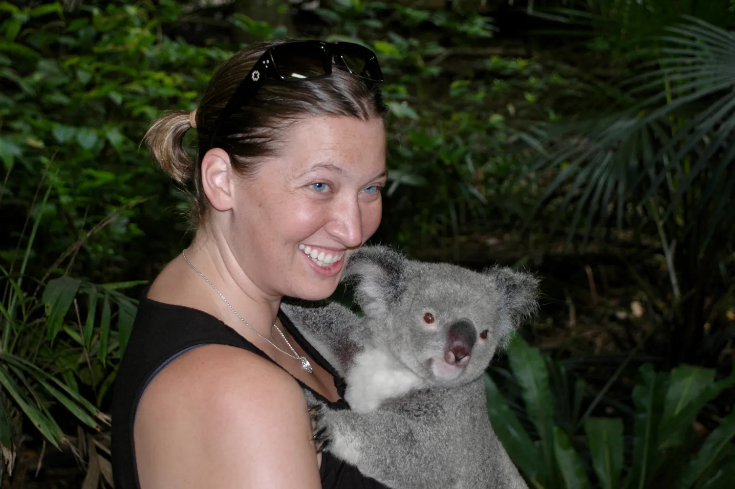 A candid portrait of a smiling woman holding a koala against a lush, green, natural background of trees and foliage. The woman, who has her hair pulled back and is wearing sunglasses on top of her head and a black sleeveless top, is looking at the camera with a bright smile. The koala has a light grey coat, a prominent white chest patch, and large, fuzzy ears, and is resting its head near the woman's shoulder.