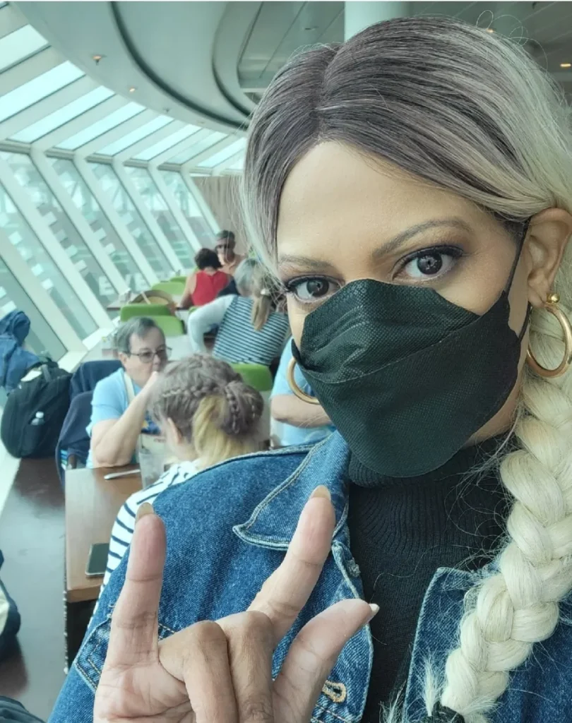A close-up selfie of a woman with a blonde-tipped braid and large gold hoop earrings. She is wearing a black face mask and a denim jacket over a black turtleneck. She holds her hand up in a "rock on" or "I love you" sign (index finger and pinky finger raised). In the background, visible through a large, curved glass window on a cruise ship or terminal, other people are seated at tables.