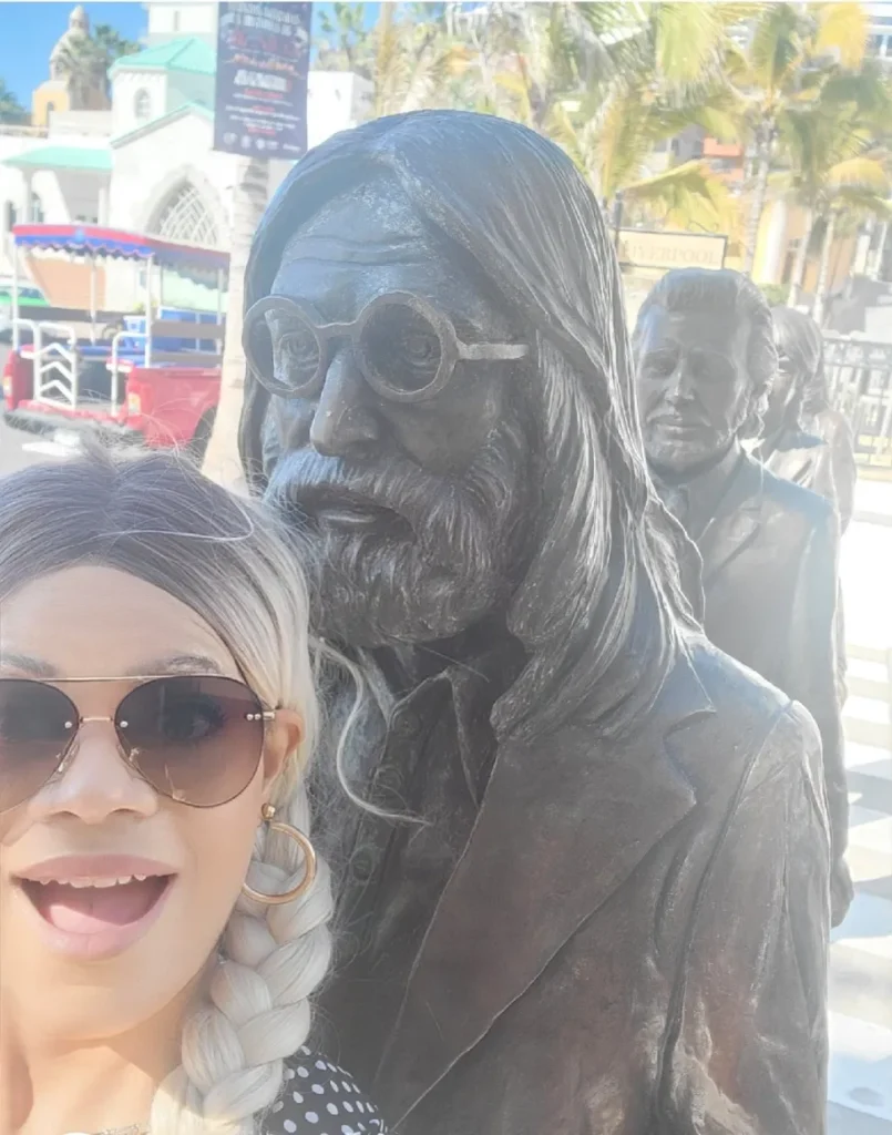 A close-up outdoor selfie of a smiling woman with a blonde-tipped braid, large gold hoop earrings, and aviator sunglasses. She is posing next to a bronze statue of John Lennon wearing his signature round glasses and beard, which is part of a line of statues of the Beatles. The statues are set against a sunny, urban background with palm trees and a historic-looking building.
