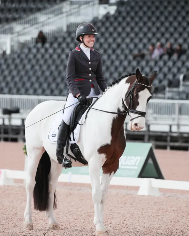 A woman in dressage competition attire—a dark jacket, white breeches, and a black helmet—smiles while sitting atop a white and brown Pinto horse. The horse has distinct brown patches over its ears and eyes. They are standing on the sand of a large indoor arena, with rows of empty seating visible in the background.