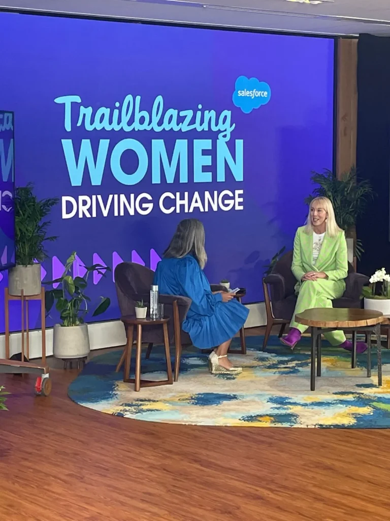 A wide shot of a stage setup for a fireside chat event. A large screen in the background displays the text "Trailblazing Women Driving Change" with the Salesforce logo. Two women are seated in upholstered armchairs facing each other. The woman on the right is wearing a bright lime green pantsuit and purple ankle boots, while the woman on the left is wearing a long blue pleated dress and gold-colored heels.
