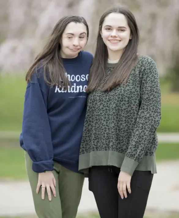 A portrait of two young women standing together outdoors. The woman on the left has long brown hair and is wearing a dark blue sweatshirt that says "Choose Kindness" and olive green pants. The woman on the right has long dark hair and is wearing a green and black leopard print sweater and black pants. They are both smiling slightly, and a soft-focus background of trees and possibly blooming pink blossoms is behind them.