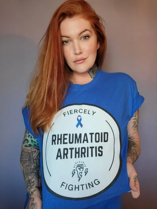 A woman with long, vibrant red hair and tattoos on her arms holds the bottom edge of a royal blue t-shirt. The t-shirt has a large white circular graphic that reads "FIERCELY RHEUMATOID ARTHRITIS FIGHTING" with a small blue awareness ribbon and a stylized fist with lightning bolts inside the circle. She is looking directly at the camera.