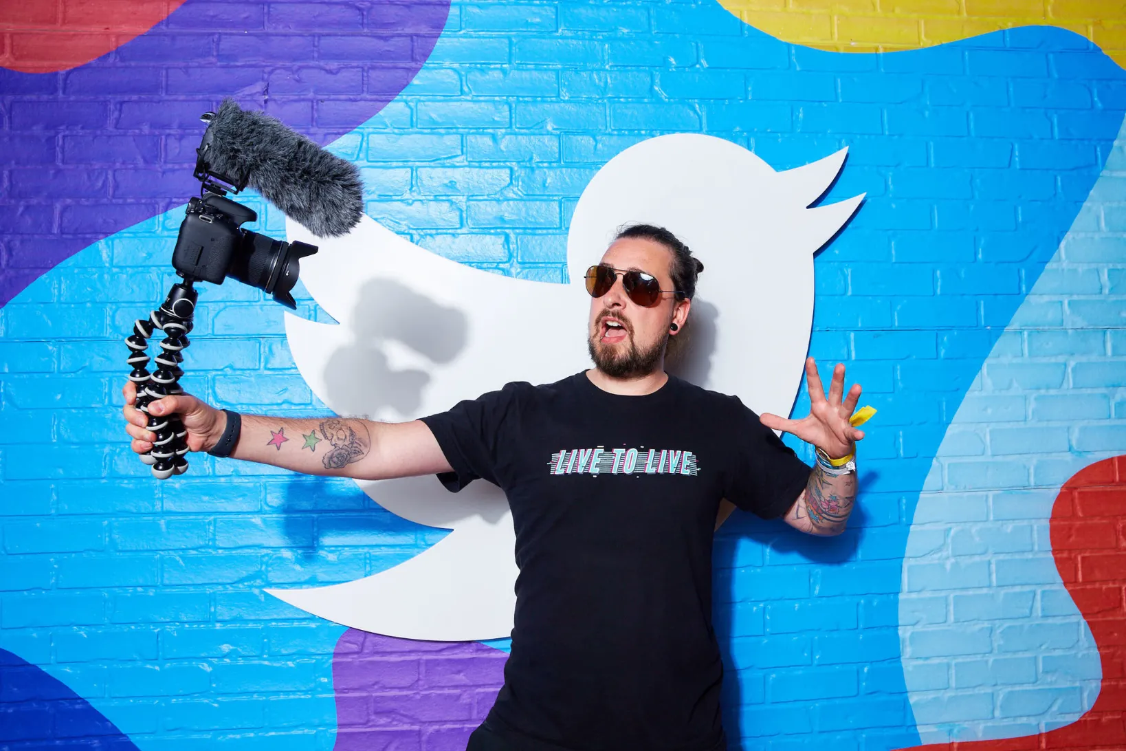 A man with a beard and sunglasses, wearing a black t-shirt that says "LIVE TO LIVE," stands in front of a brightly colored brick wall painted with a large white Twitter (X) bird logo. He is holding a camera mounted on a small, flexible tripod (a Gorillapod) with a microphone attachment in his outstretched right hand, seemingly recording himself or a video. He has tattoos on both arms.
