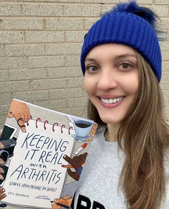 A close-up, outdoor portrait of a young woman with long brown hair, wearing a bright blue knit beanie with a pom-pom and a grey sweatshirt. She is smiling directly at the camera while holding a book titled, "Keeping It Real with Arthritis: Stories from Around the World" by Effie Kolipoulos. The book's cover features a spiral notebook design with illustrations of hands, pencils, and a coffee cup. A brick wall is visible in the background.