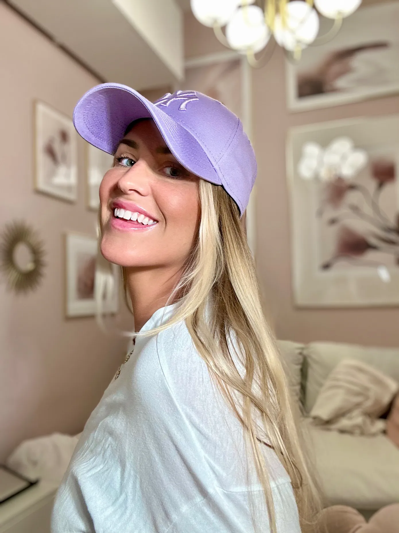 A smiling blonde woman wearing a lavender baseball cap is seated indoors, looking over her shoulder at the camera. The background is a soft pink room with white framed pictures and a fluffy white couch, creating a cozy and warm atmosphere.
